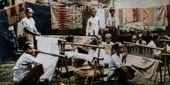 buat gambar foto ukuran 16:9. Konsep foto: Seorang gadis Indonesia menggunakan kain dodot ditutupi selendang warna kuning sedang merentangkan kain putih dengan motif batik belum jadi di gawangan. Masa itu era tahun 1930-an.