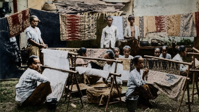 buat gambar foto ukuran 16:9. Konsep foto: Seorang gadis Indonesia menggunakan kain dodot ditutupi selendang warna kuning sedang merentangkan kain putih dengan motif batik belum jadi di gawangan. Masa itu era tahun 1930-an.